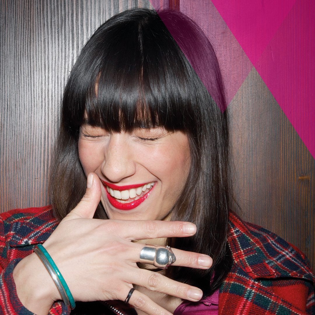Person with long dark hair wearing a red plaid jacket, showing rings and bracelets on their hand, standing against a wooden background.