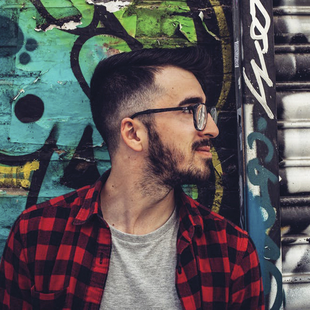 A man with glasses and a beard, wearing a red checkered shirt, standing against a vibrant graffiti wall.