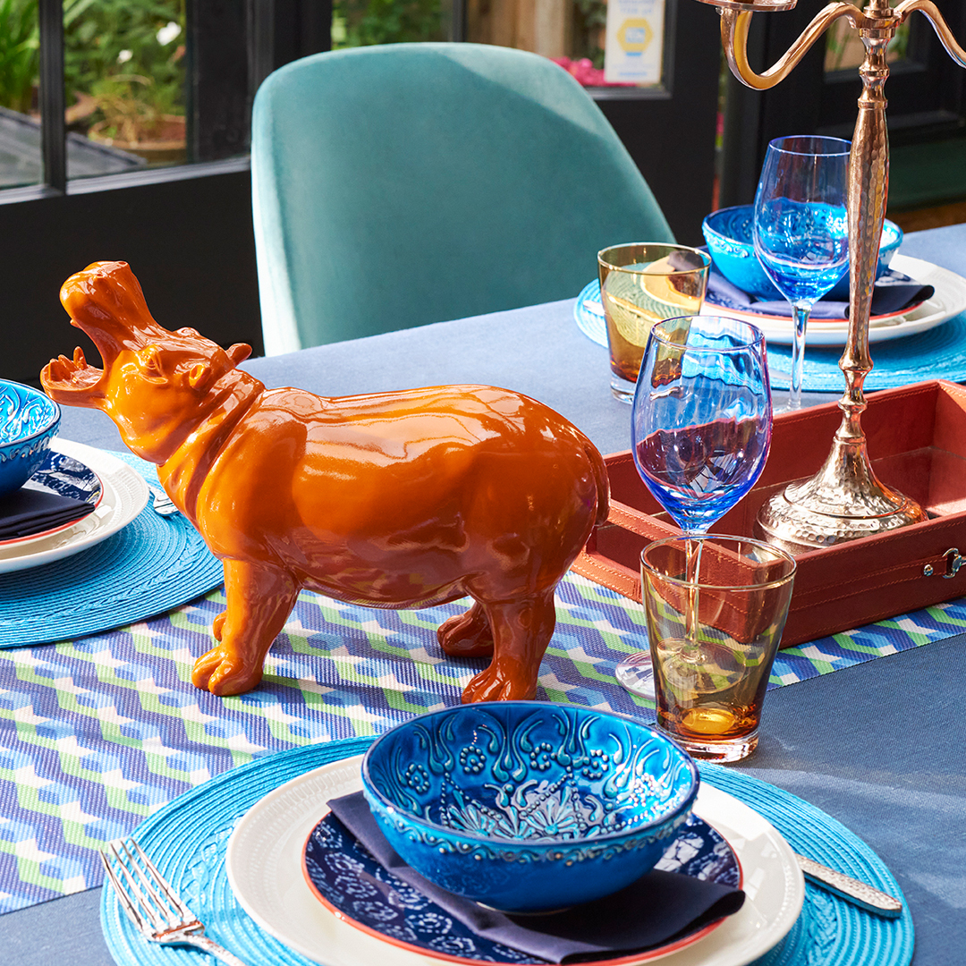 A vibrant dining table set with blue plates, colorful glasses, and a striking orange hippo sculpture.