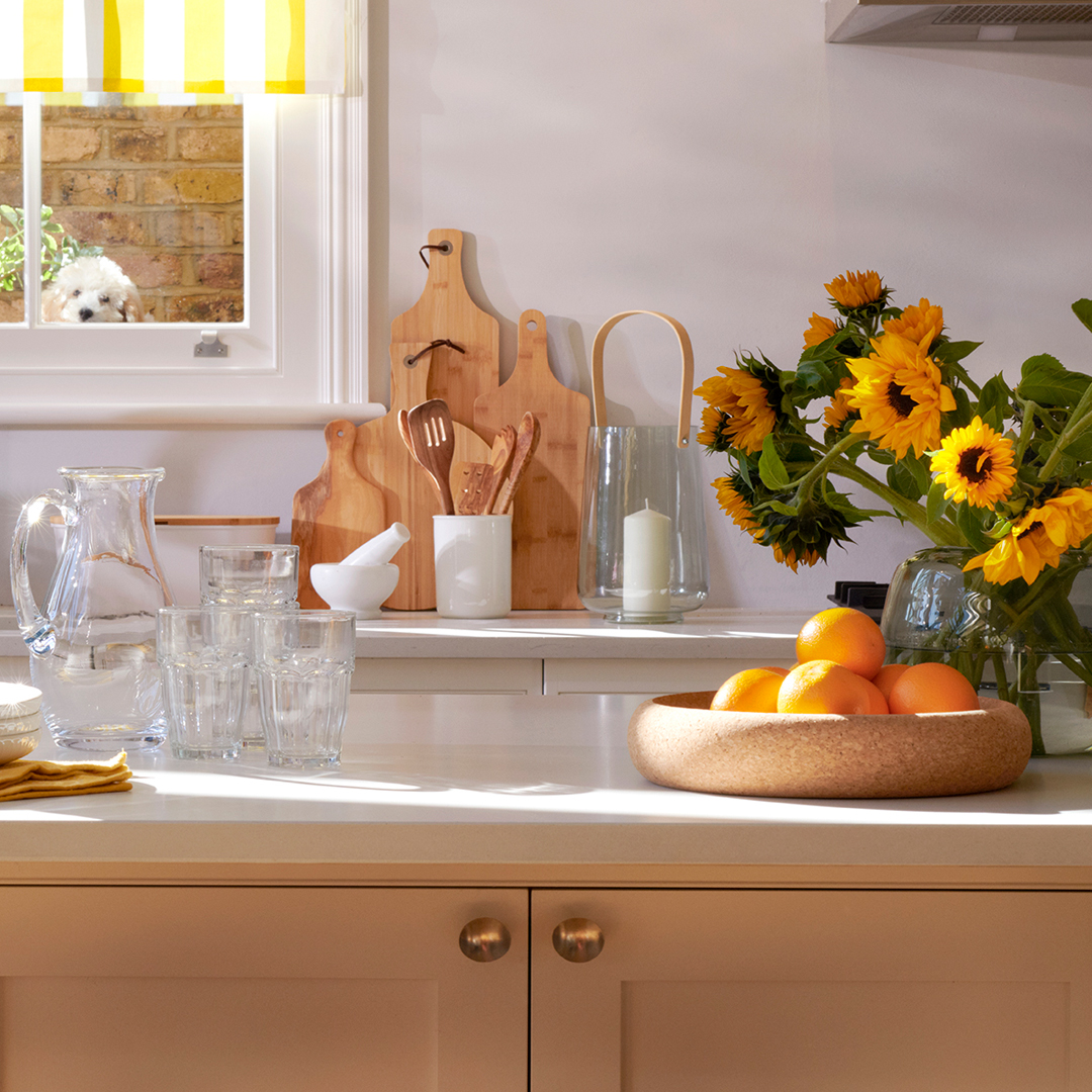 A sunny kitchen counter with wooden cutting boards, a pitcher of water, glasses, a bowl of oranges, and a vase of sunflowers. A dog can be seen peeking through the window.