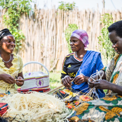 A group of three women sitting together, smiling and weaving baskets in a sunny outdoor setting.