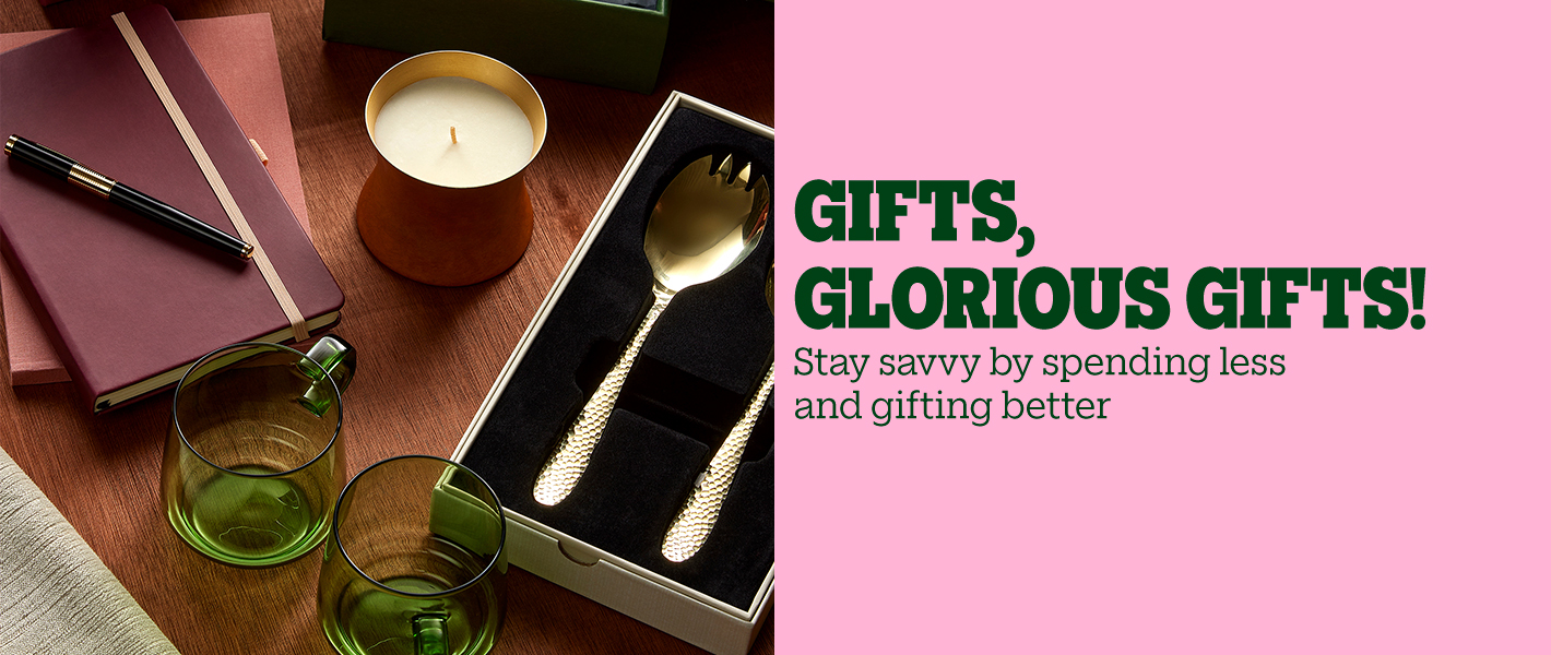 Gift items including notebooks, a pen, a candle, green glass cups, and a boxed gold serving set on a wooden table, with the text "Gifts, glorious gifts! Stay savvy by spending less and gifting better.