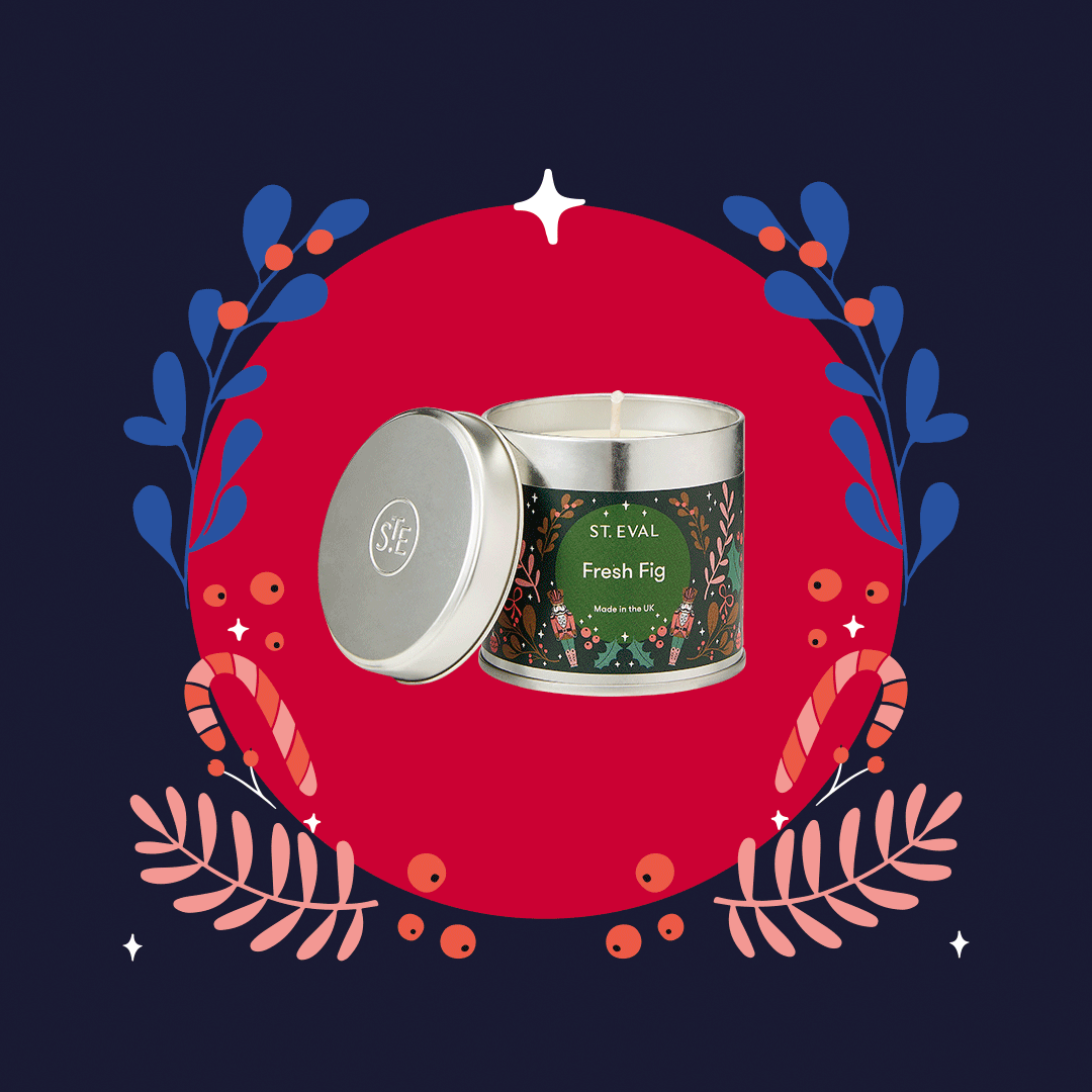 A silver tin candle with a "Fresh Fig" label from St. Eval, set against a festive red background with decorative foliage and candy canes. The candle is made in the UK.