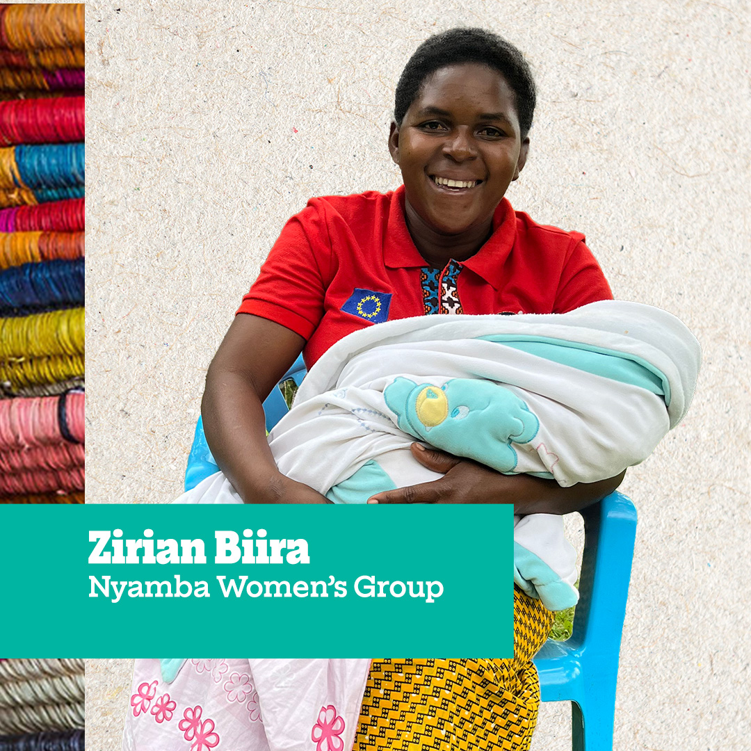 A woman named Zirian Biira from the Nyamba Women’s Group, smiling while holding a baby wrapped in a blanket, sitting on a blue chair.