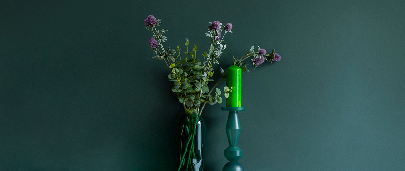 Green glass vase with eucalyptus stems and purple flowers beside a green candle on a modern candle holder, set against a dark green wall.