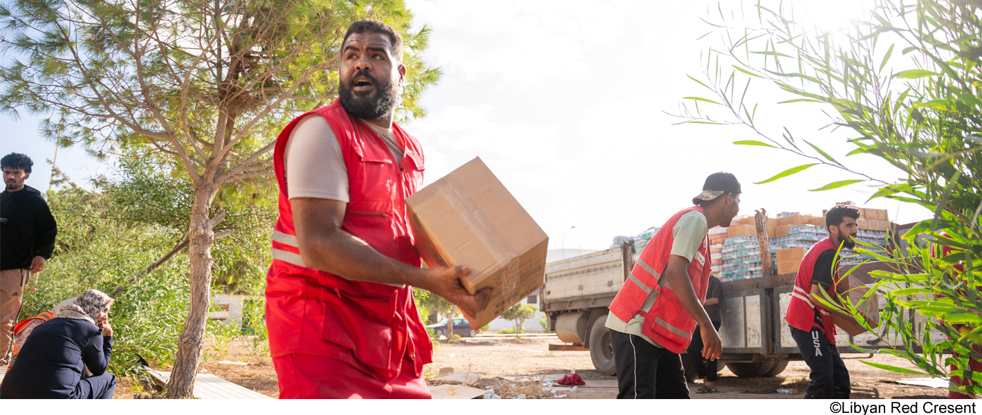 Volunteers in red vests unloading supplies from a truck, with one carrying a box while others assist in the background.