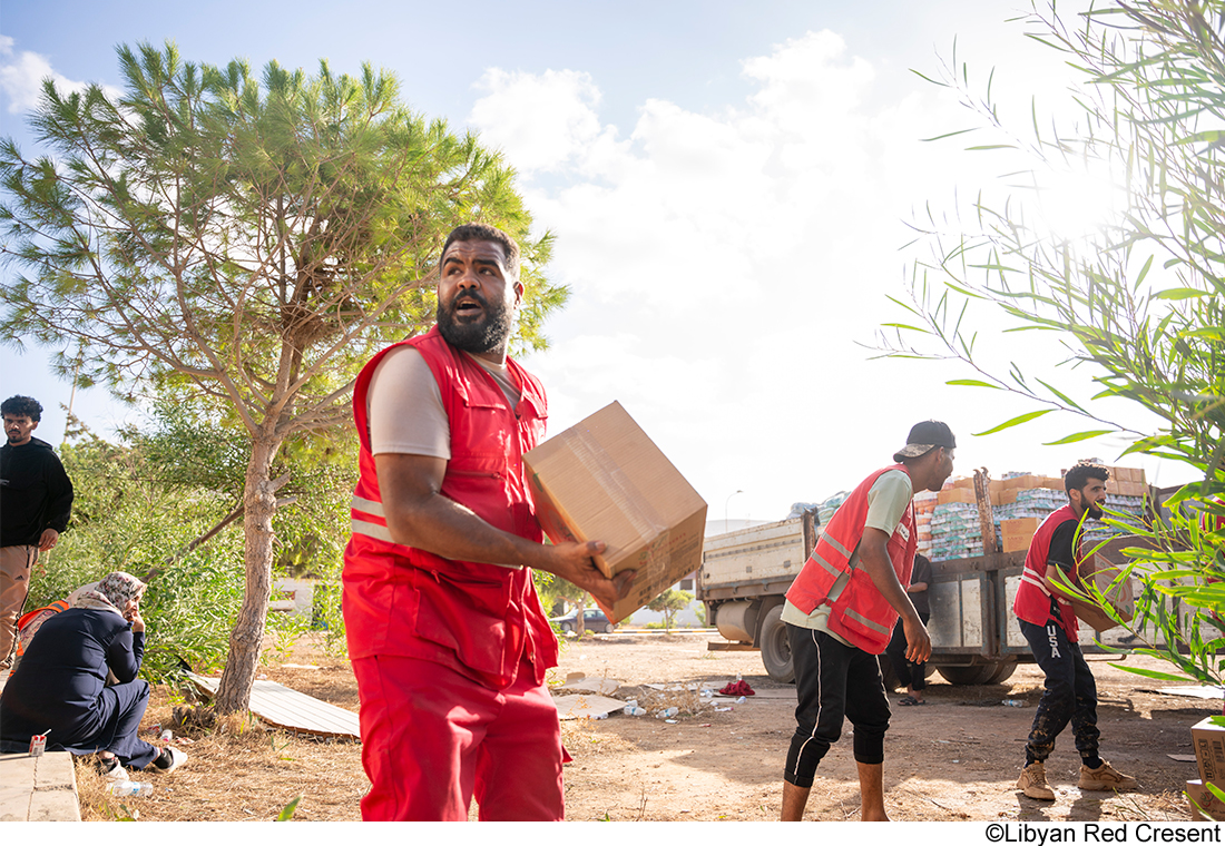Volunteers in red vests unloading supplies from a truck, with one carrying a box while others assist in the background.