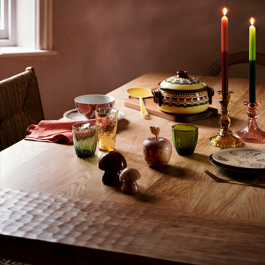 Cozy dining table setting with colorful glassware, ceramic dishes, candles, and decorative mushrooms on a wooden table.