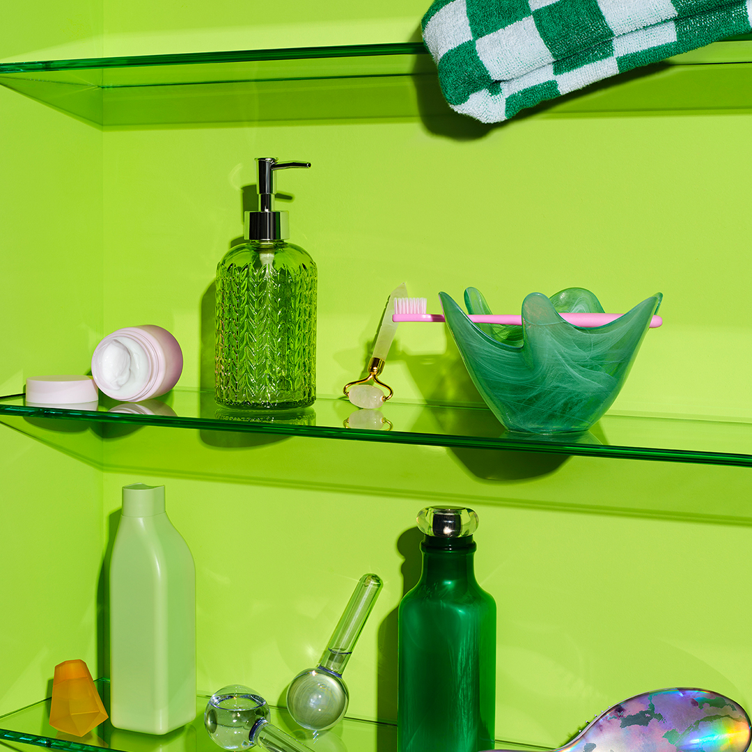 Bright green bathroom shelves with skincare products, a toothbrush in a green bowl, a green towel, glass soap dispenser, and other toiletries.