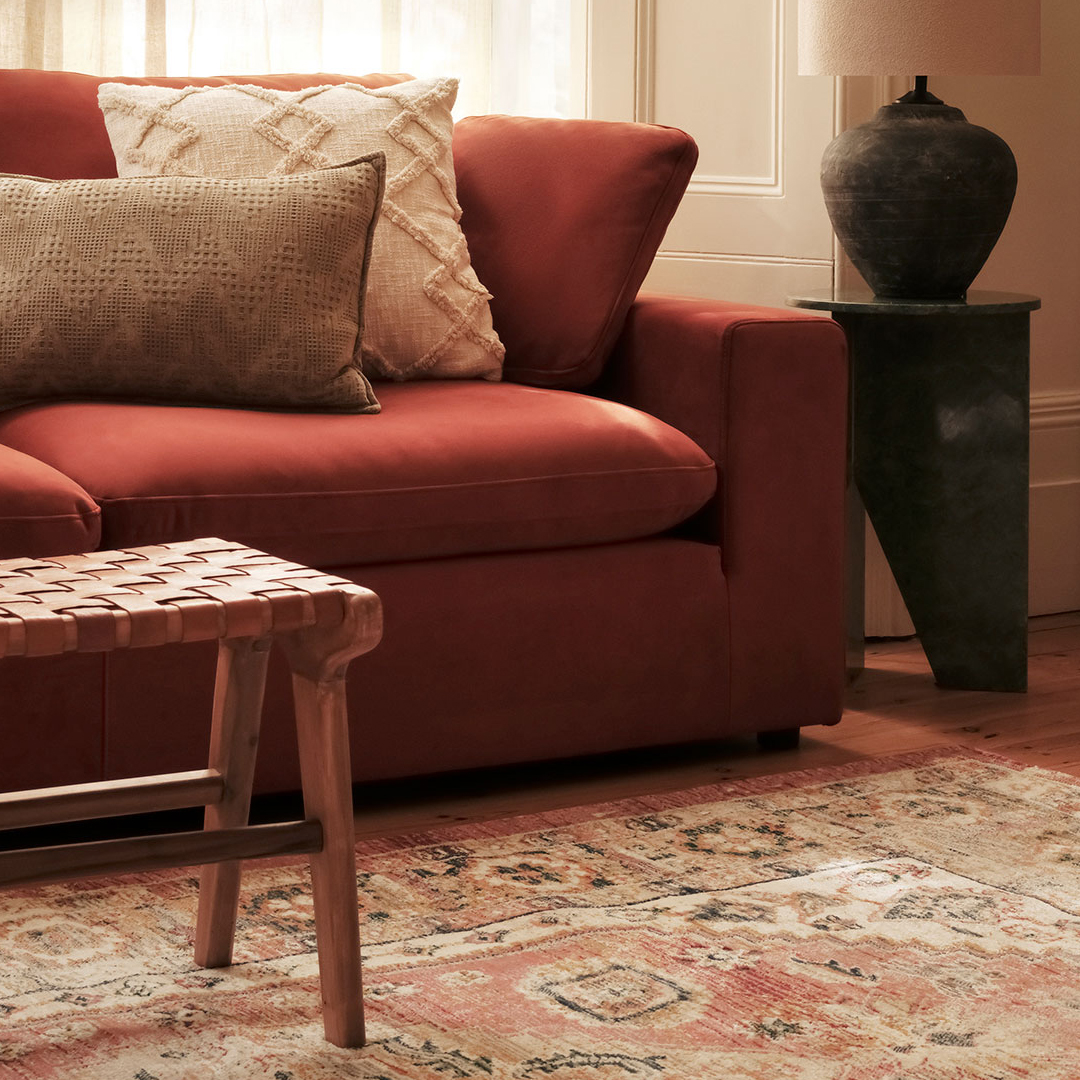 A cozy living room with a rust sofa, textured pillows, woven stool, patterned rug, and dark ceramic lamp on a green side table.