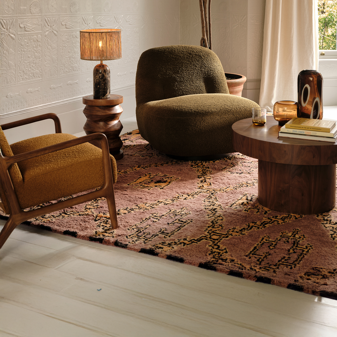 Stylish living room with a textured olive chair, mid-century wood armchair, sculptural side table with lamp, round coffee table, and patterned rug.