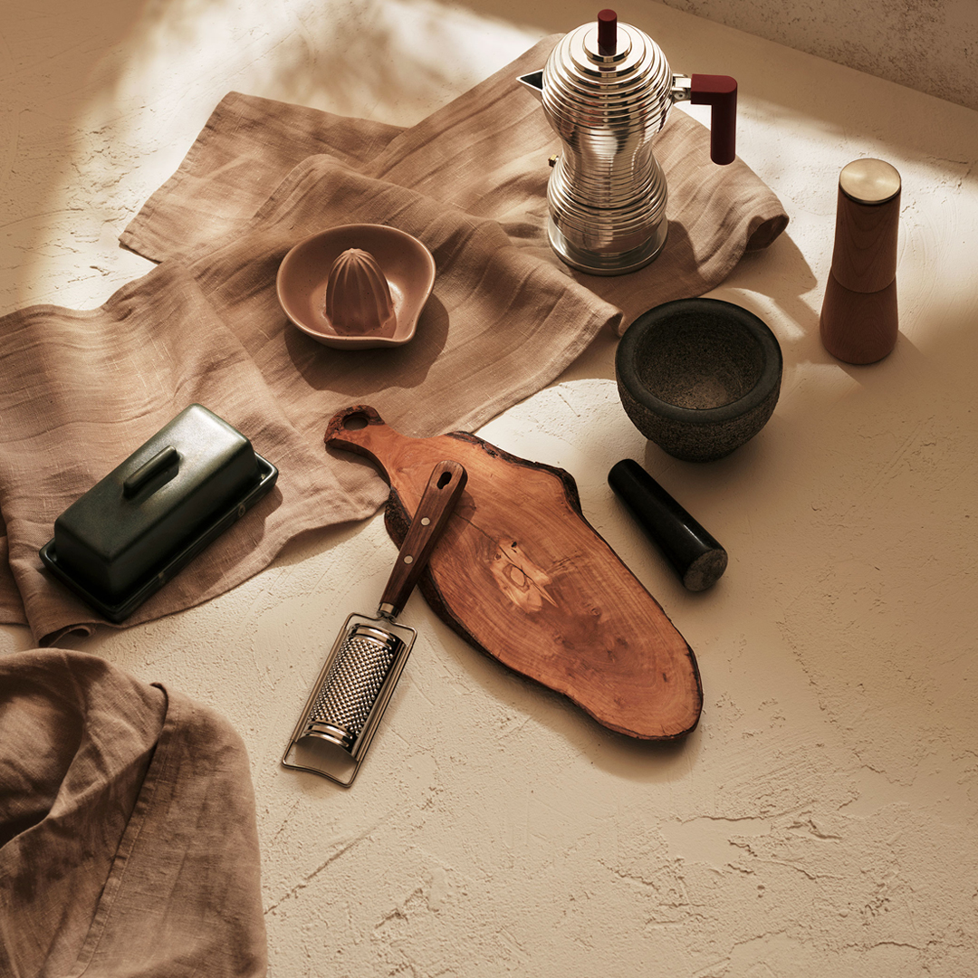 A flat lay of rustic kitchen tools including a wooden cutting board, cheese grater, citrus juicer, butter dish, moka pot, mortar and pestle, and a pepper grinder on a textured surface with neutral-toned linens.