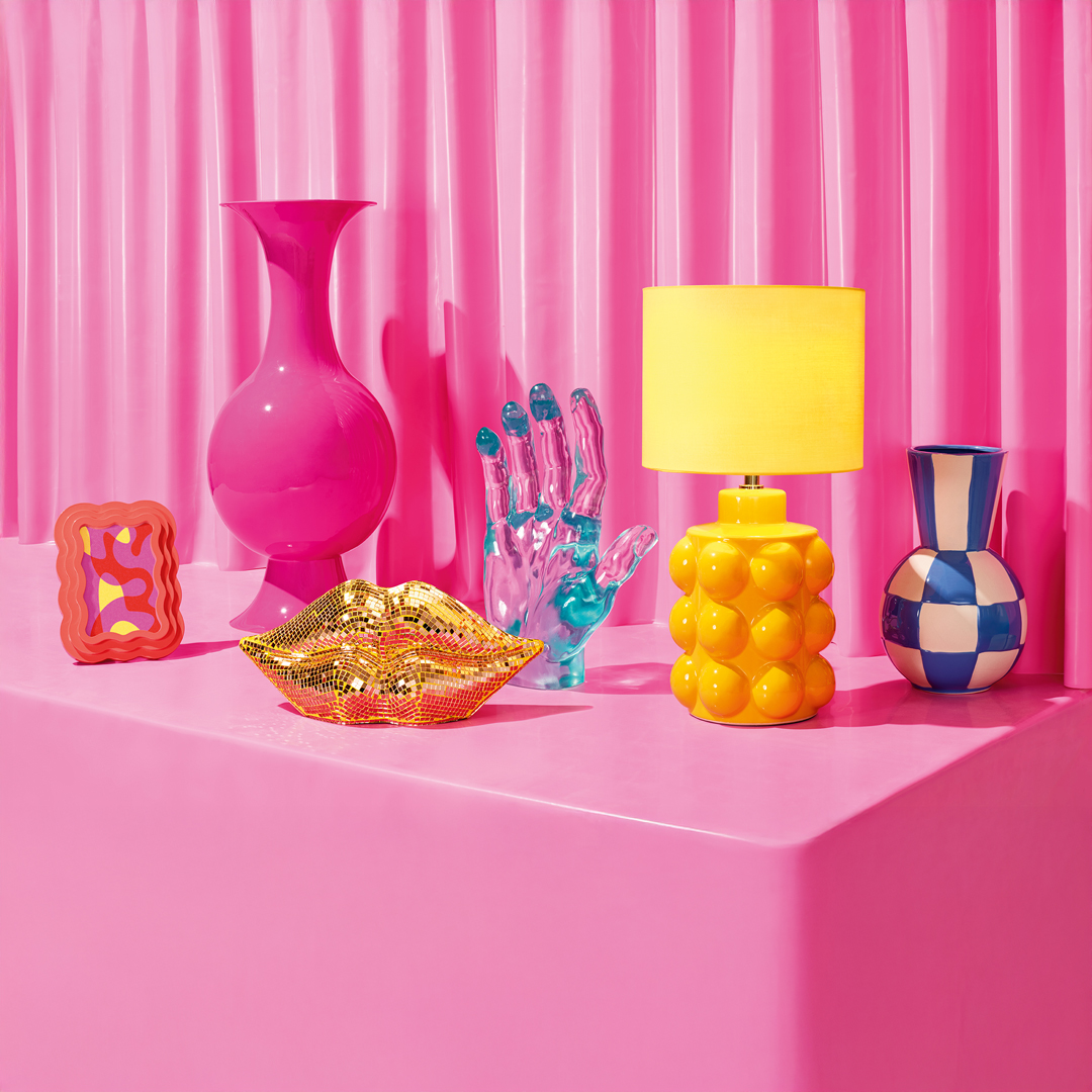 Bright pink vase, orange frame, gold lips sculpture, transparent blue hand, yellow lamp, blue and white checkered vase on pink surface.