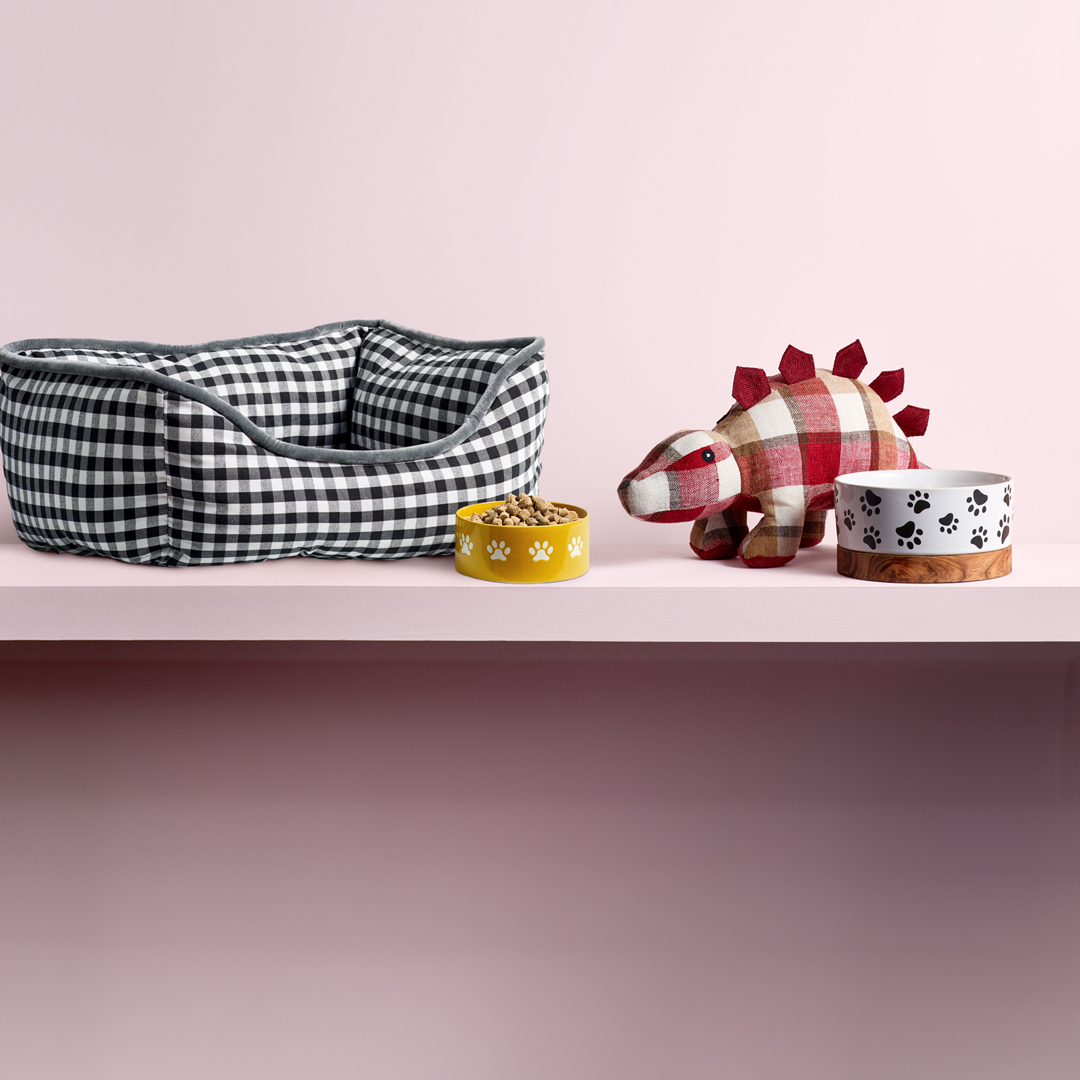 Black and white checkered pet bed, yellow and white paw-print food bowls, and a red plaid dinosaur toy on a pink surface.