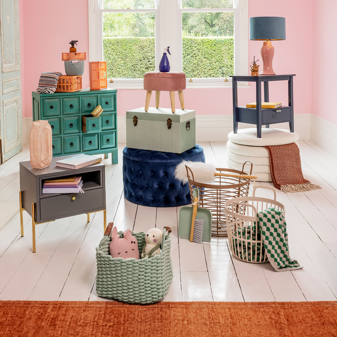 Colorful room with various storage baskets, drawers, stools, and decorative items arranged on a white wooden floor with pink walls and large windows.