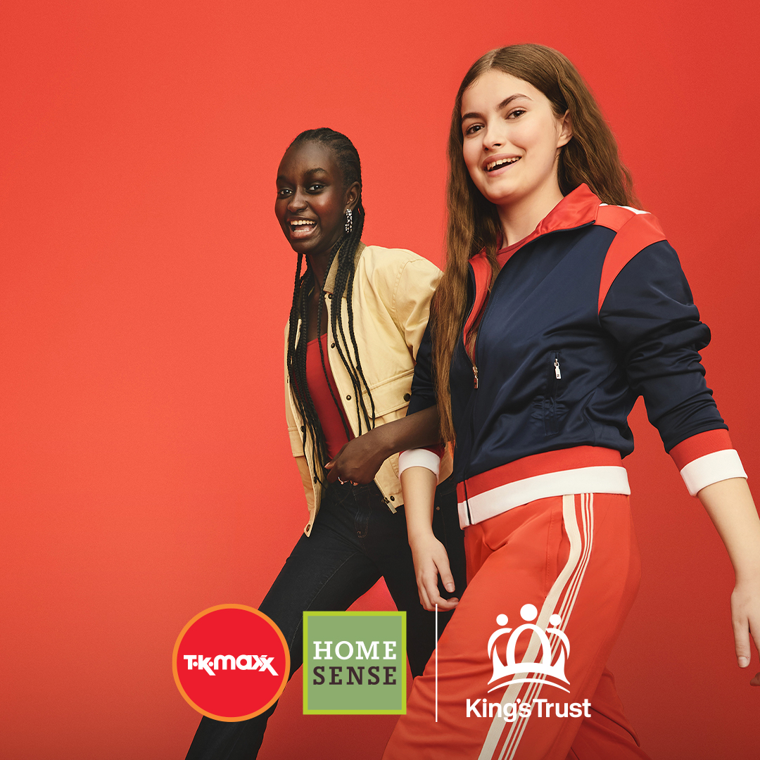 Two girls walking with their arms linked smile as they look towards the camera.