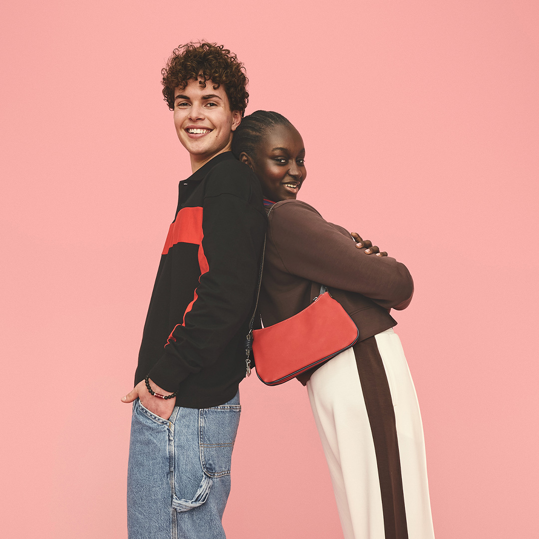 A boy and girl lean against each other standing back-to-back and smile at the camera.