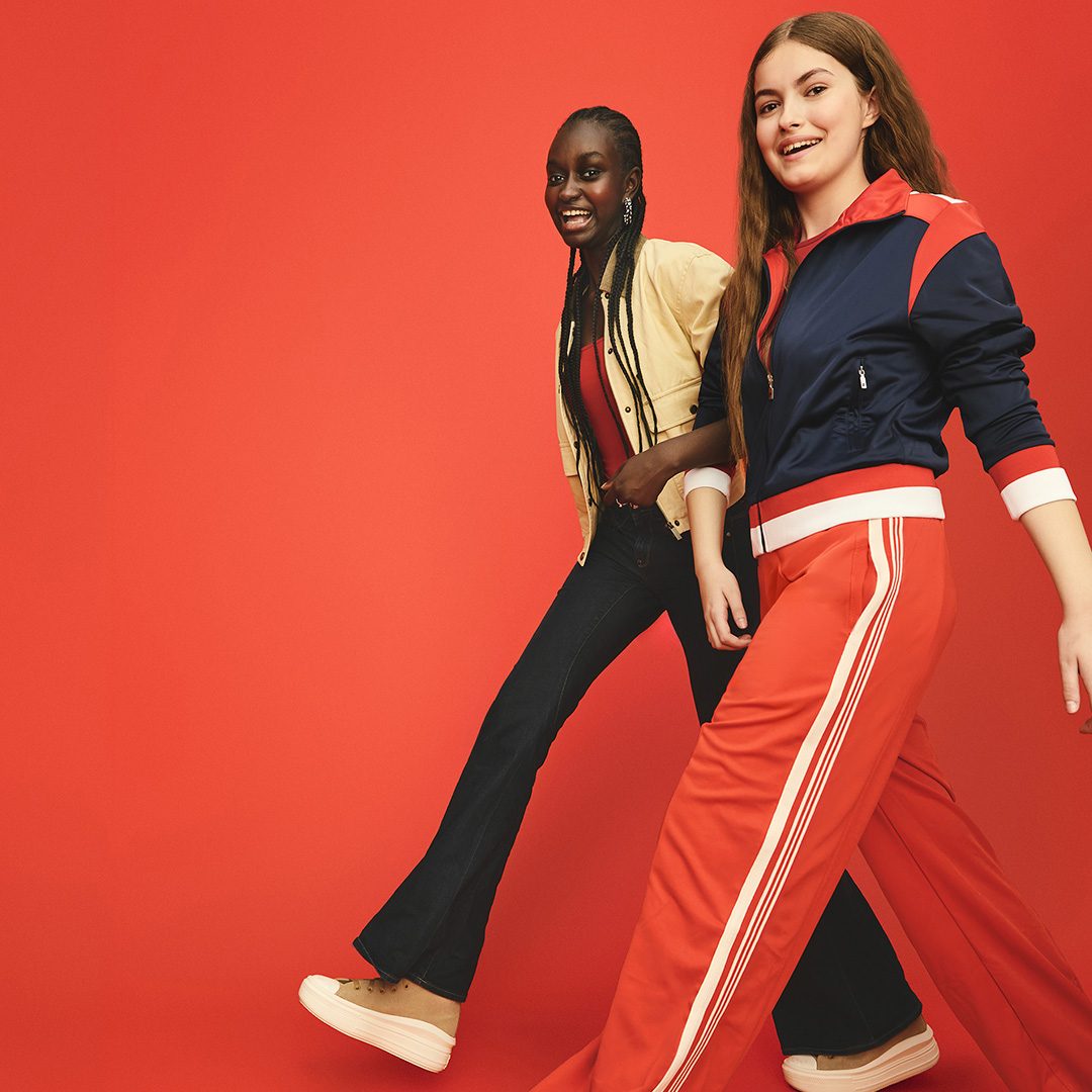  Two girls stride in step with each other while smiling at the camera.