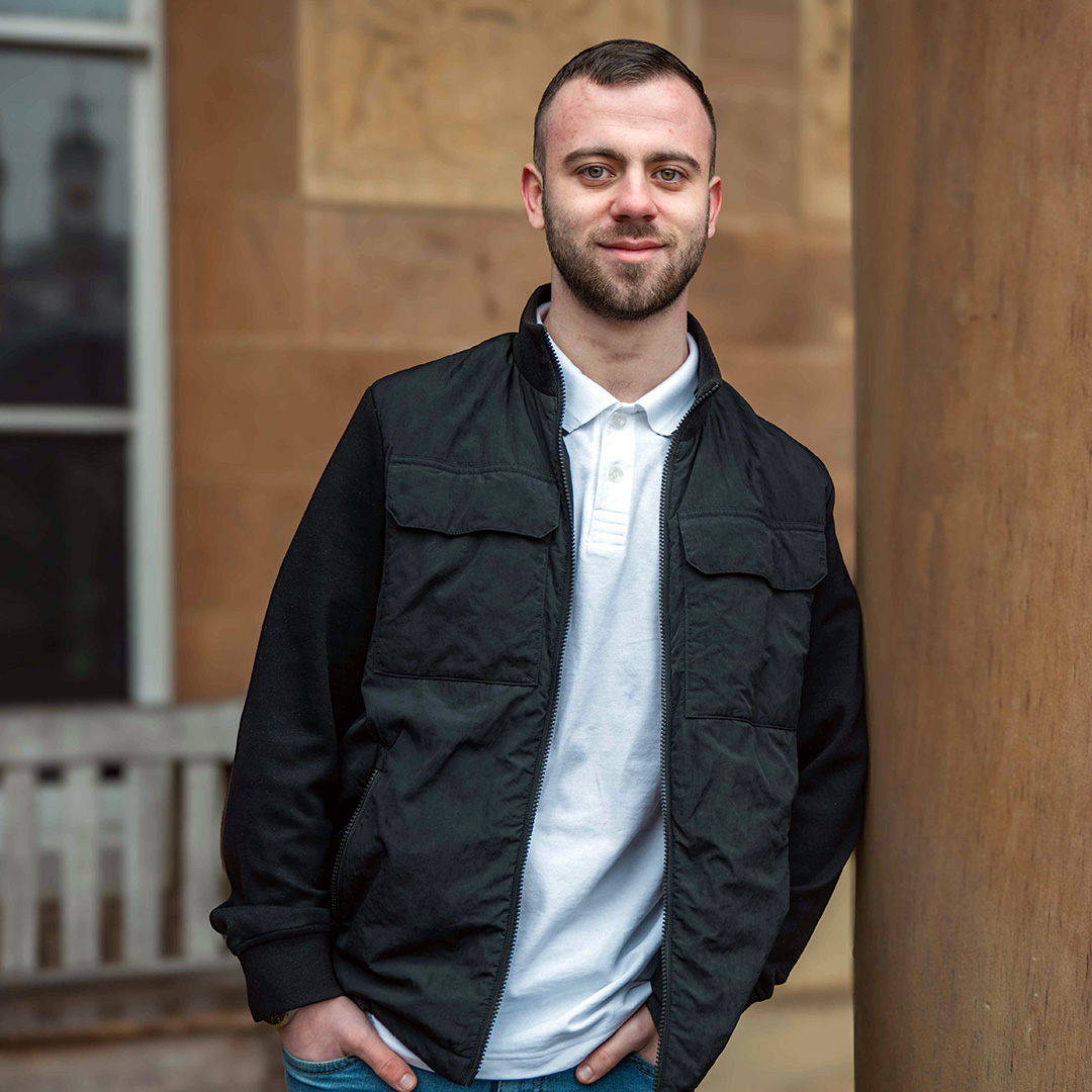Patrick stands in front of a sandstone building with his hands in his pockets.